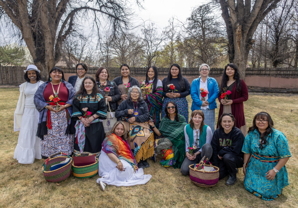 Large group of colorfully dressed women, some in traditional clothing, standing and sitting outside. Doula graduation ceremony at Tewa Women United campus, 2025